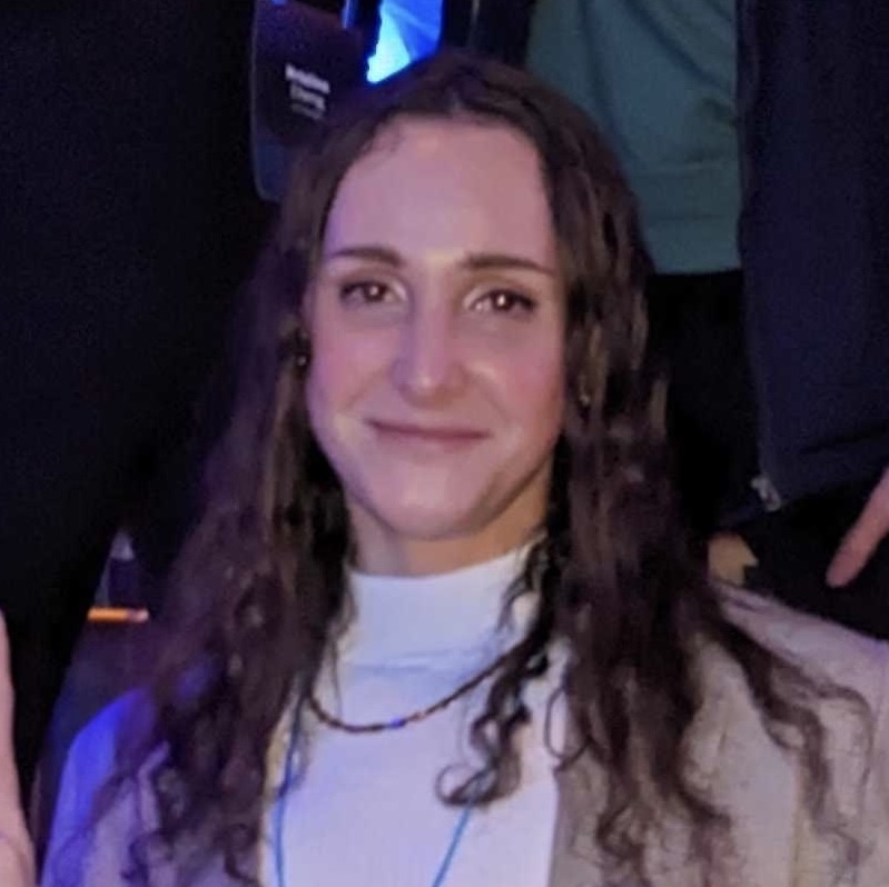 A headshot of me, Lily Roberts.  I am at a work event in a music venue, wearing a checkered blazer and a white ribbed turtleneck.